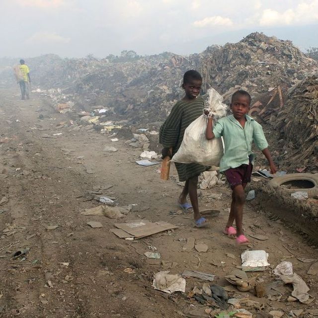 Kinder laufen mit Müllsäcken auf dem Rücken an einem Müllberg vorbei, im Hintergrund ist giftiger Rauch zu sehen.