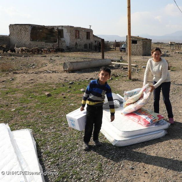 Ein Junge trägt eine Kiste mit der Aufschrift "UNHCR", eine Frau steht im Hintergrund und hebt zwei Säcke von einer Matratze auf.