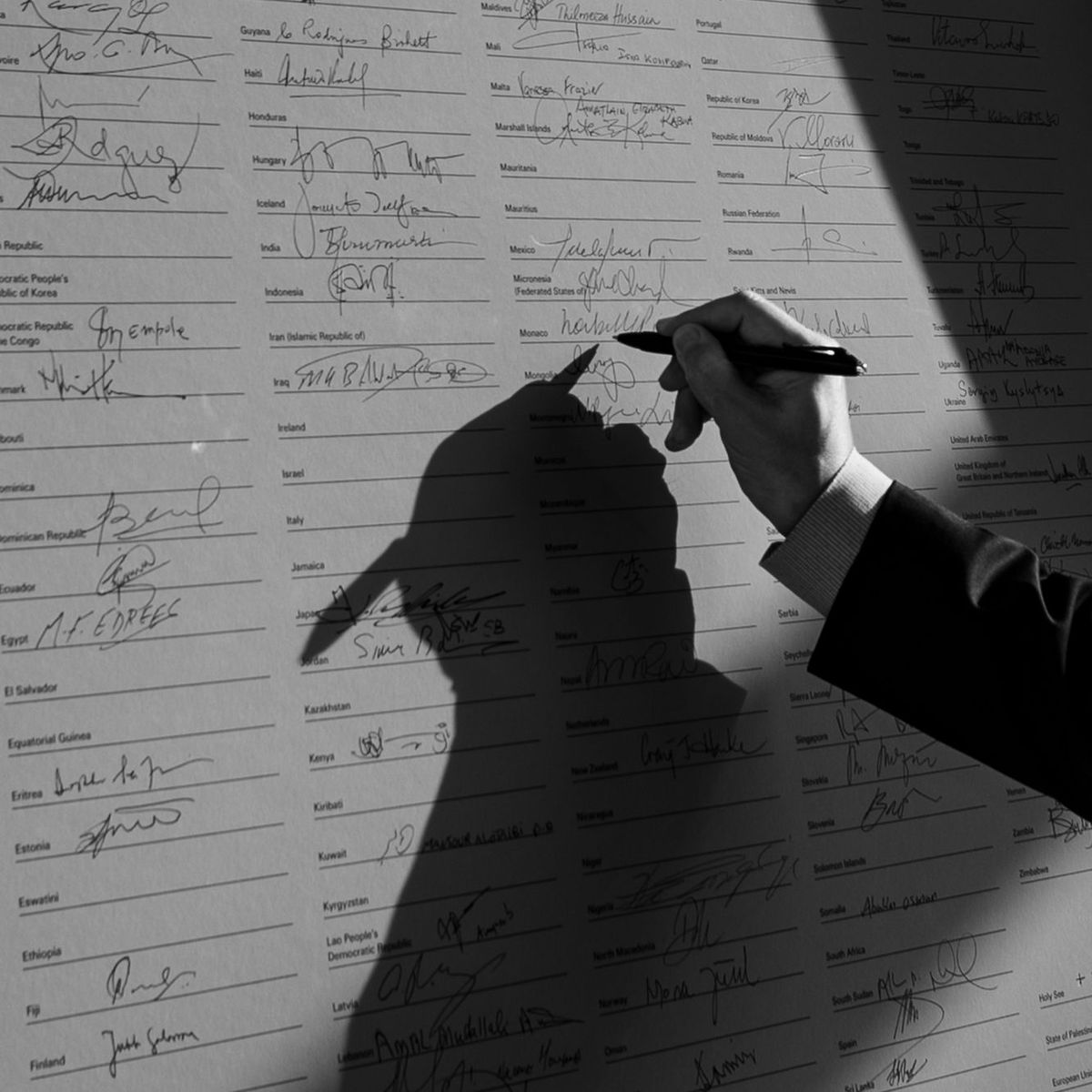 Ein Schwarz-Weiß-Foto zeigt eine Hand, die gerade ihre Unterschrift auf eine große Liste von anderen Unterschriften setzt.