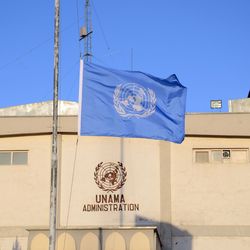 Die Flagge der Vereinten Nationen auf Halbmast am Hauptquartier der UN-Unterstützungsmission in Afghanistan (UNAMA) in Kabul. Vor einem sandfarbenen Gebäude mit dem UNAMA-Logo weht die blaue UN-Flagee auf Halbmast.