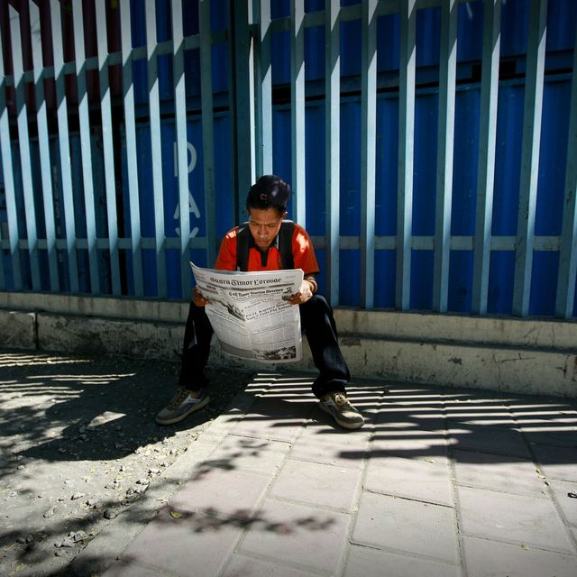 Ein junger Mann sitzt vor einer blauen Metallgitterbarriere alleine und liest gedankenverloren die Zeitung.