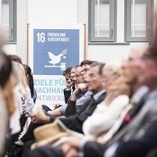 Menschen lauschen einem Vortrag. Im Hintergrund steht ein Banner zu SDG 16.