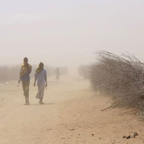 Zwei Menschen laufen auf einer sandigen Straße durch einen Sandsturm, sie haben Tücher um den Kopf gewickelt