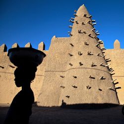 Ein sandfarbenes kuppelförmiges und oben spitz zulaufendes Gebäude, davor der Schattenriss einer Frau.