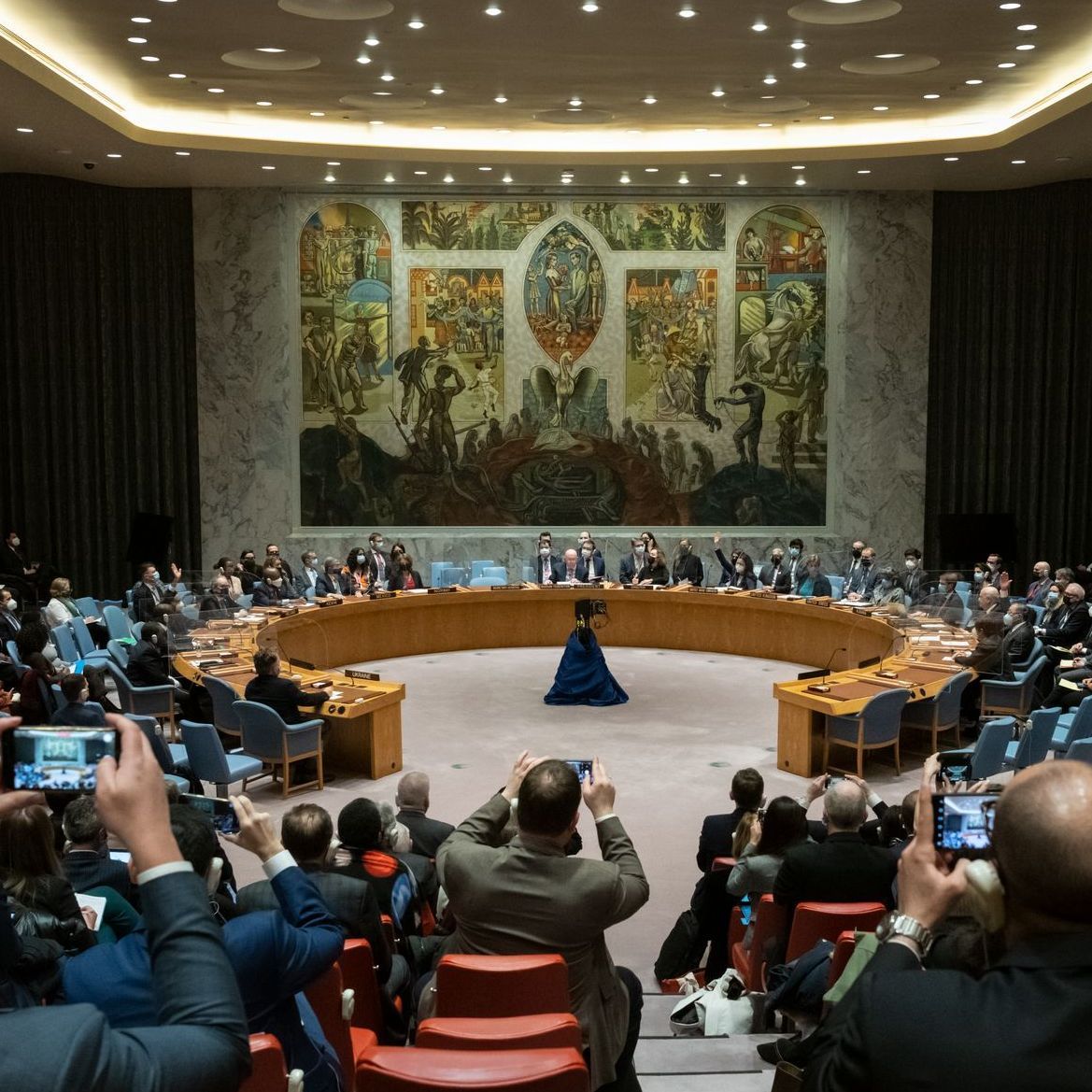 Blick in den UN-Sicherheitsrat: Um einen runden Tisch herum sitzen einige Menschen, Fotografen dokumentieren das Geschehen.