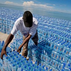 Ein Mann sortiert hunderte Wasserflaschen, im Hintergrund sieht man das Meer.
