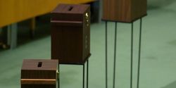Blick auf drei Wahlurnen aus Holz, die in der Generalversammlungshalle aufgestellt sind. 